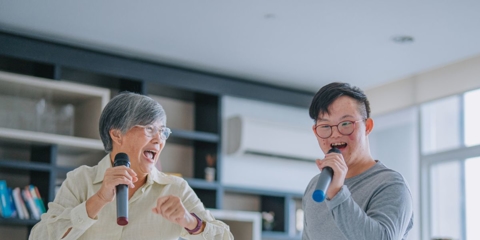 Photo of an older woman and younger man singing into microphones in their living room.