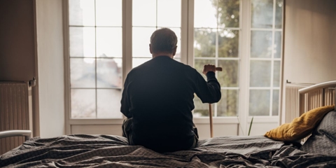 Photo of a senior man sitting on a bed in front of a large window, resting his hand on a cane.