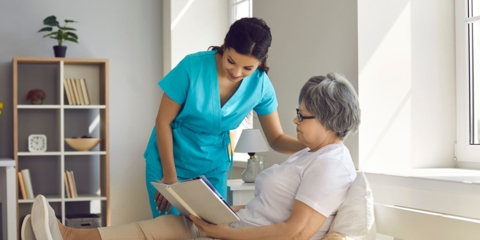 Photo of an in-home care attendant in blue reading a book on an older woman's lap in a home setting.
