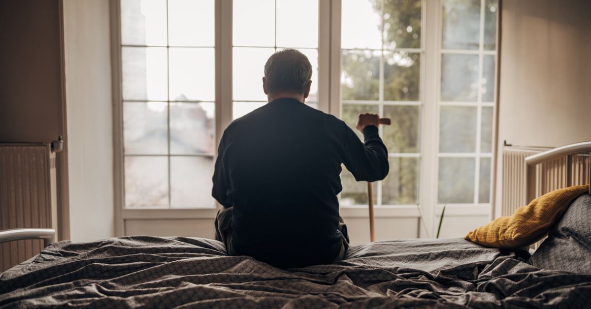 Photo of a senior man sitting on a bed in front of a large window, resting his hand on a cane.