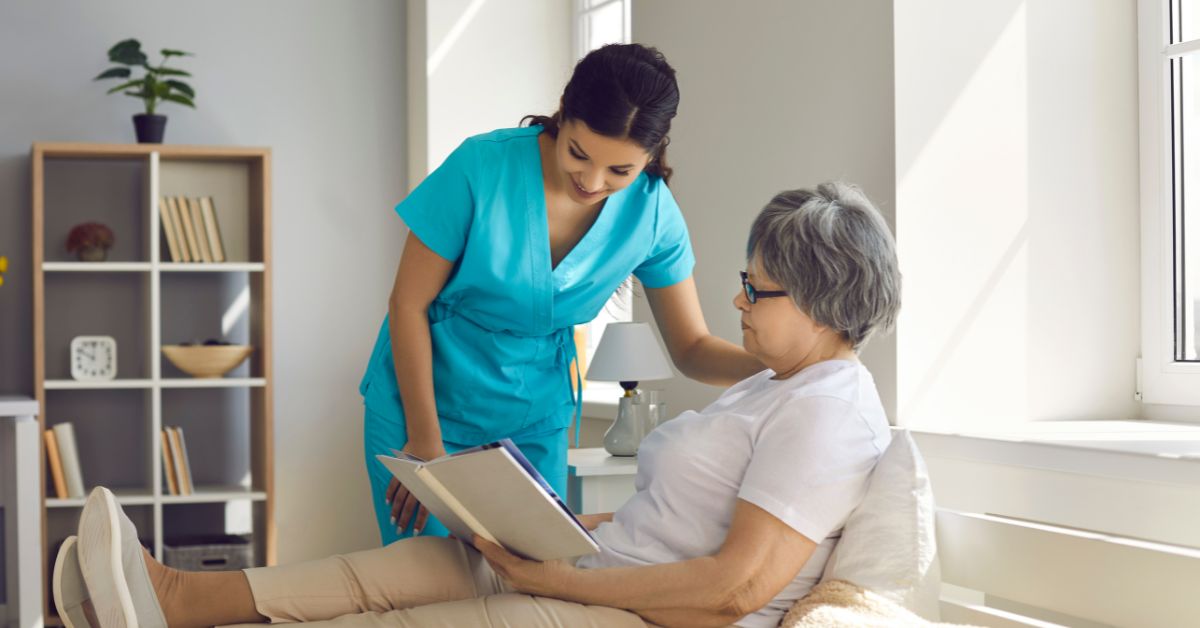 Photo of an in-home care attendant in blue reading a book on an older woman's lap in a home setting.